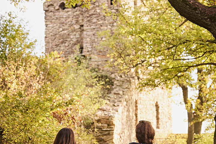 Zwei Frauen wandern mit ihren Rucksäcken und Wanderoutfit auf die Ruine Rossel in Rüdesheim am Rhein zu.